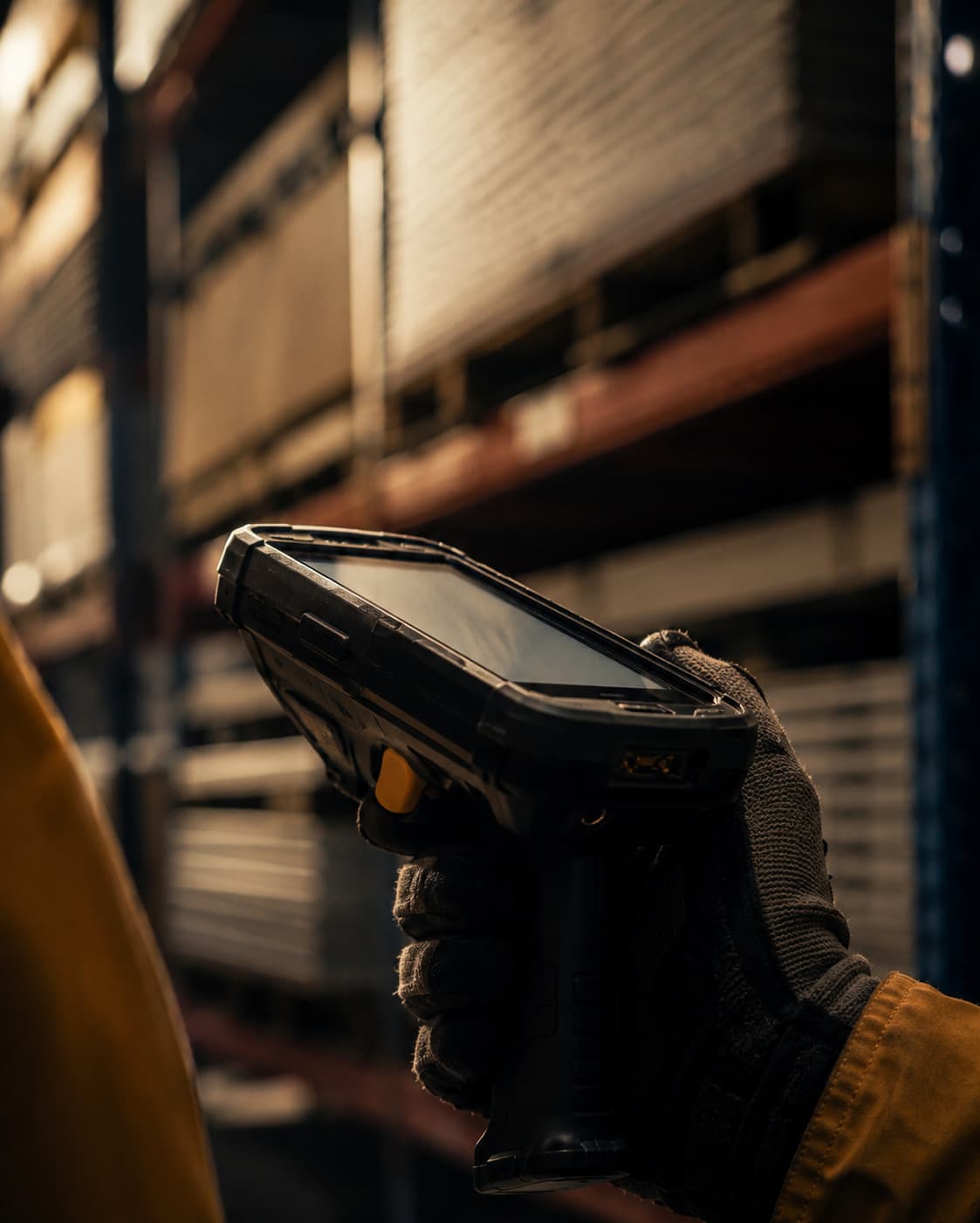 Gloved hand holding a rugged RF barcode scanner in front of steel warehouse racking.